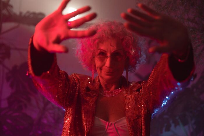 Older woman wearing a sparkly jacket and sunglasses dancing with hands raised in colorful lighting at a wild party scene.