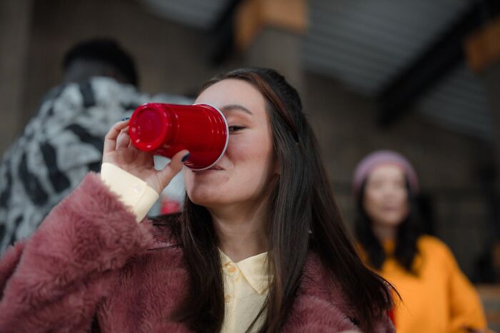 Young woman in a pink coat drinking from a red cup at a social gathering, illustrating terrifying statistics about sleep issues.