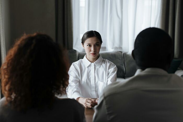 A woman in a white shirt, sitting across from a couple, looking seriously. This highlights family secrets parents told their kids.
