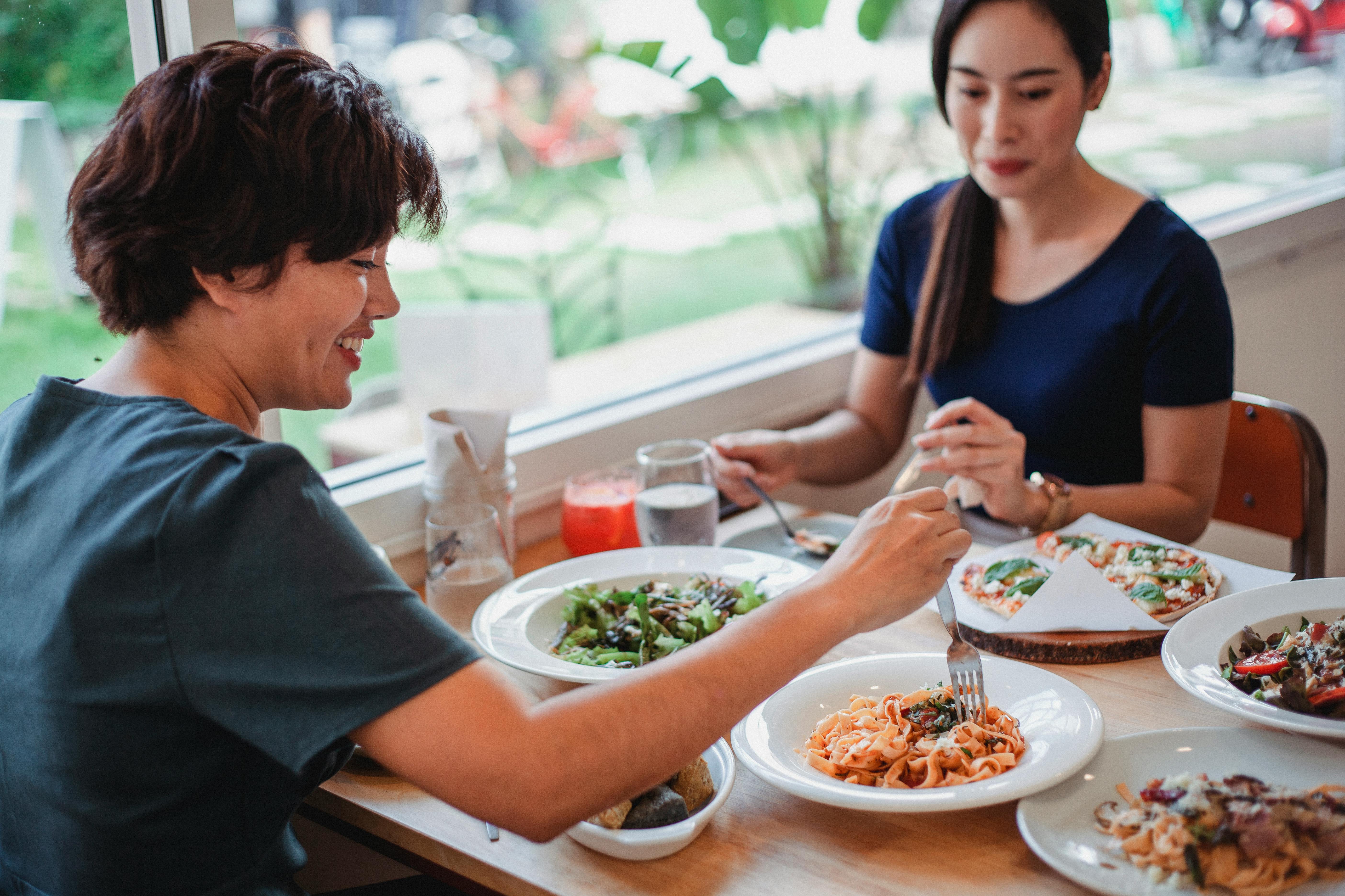 Two women enjoying a meal together at a restaurant, sharing pasta, salad, and pizza in a casual setting for social dilemma game.