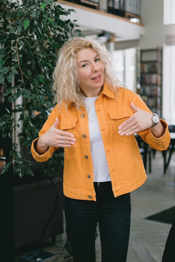 Woman in yellow jacket explaining dark family secrets indoors with plants and bookshelves in the background
