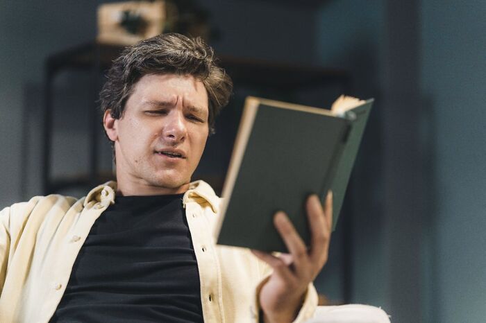 Man sitting indoors, looking confused while reading a book about terrifying statistics that might cause sleep troubles.