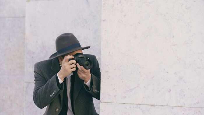 A person in a fedora and trench coat peeks from behind a wall, holding a camera. Capturing family secrets.