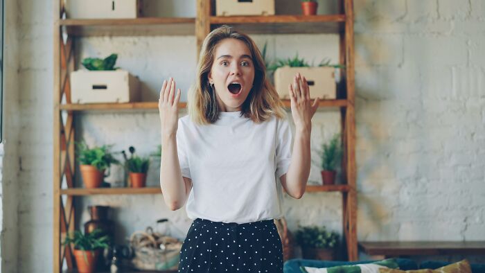A surprised young woman with short blonde hair and a white t-shirt, hands raised in disbelief about the Mandela Effect.