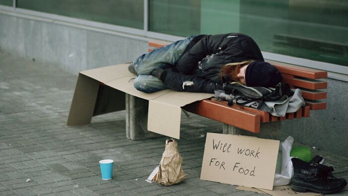 Homeless person sleeping on a bench with a sign saying will work for food, illustrating unexpected and bizarre ways people passed away.