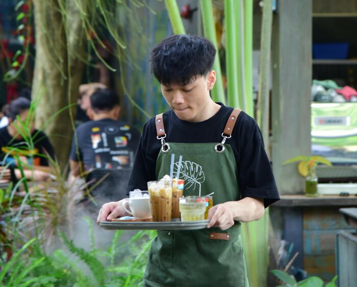 A young man in a green apron carries a tray with drinks, amidst lush greenery. He might have heard some wildest rumors.