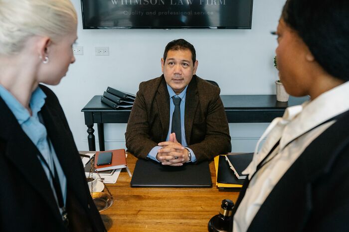 A male lawyer sits at a desk, flanked by two women, possibly discussing workplace incidents or legal matters.