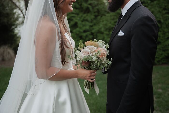 Bride in white dress holding bouquet talking to groom in black suit outdoors, representing amazing stories about their moms.