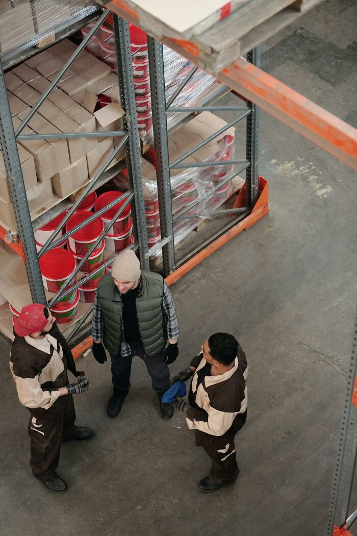 Warehouse employees discussing work near shelves with stacked boxes and paint buckets in an industrial setting.