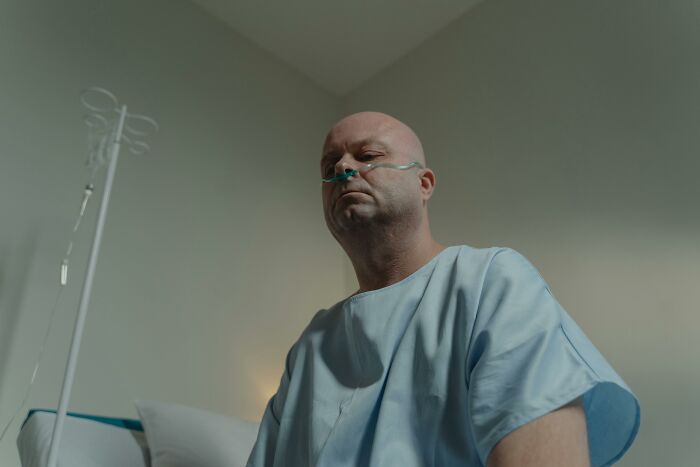 A bald patient with a nasal cannula in a hospital gown, seen from a low angle, reflecting the lasting impact of workplace incidents.
