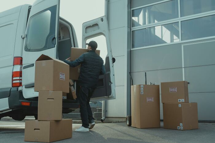 A man loads brown boxes into a white delivery van, surrounded by more boxes. Workplace Incidents can occur during such tasks.