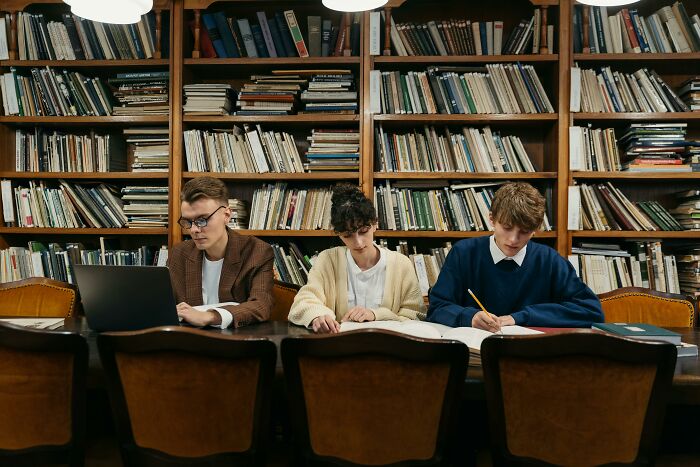 Three young people studying general knowledge with books and a laptop in a library filled with shelves of books.