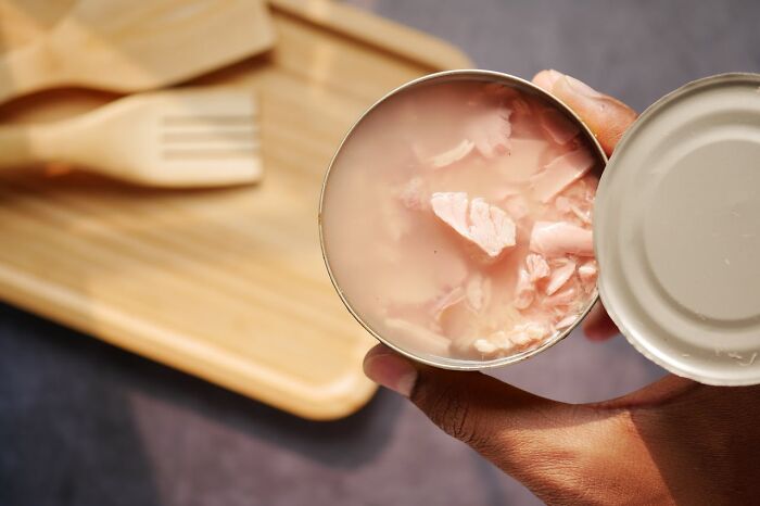 Opened can of pink tuna held by hand above wooden cutting board with wooden kitchen utensils nearby