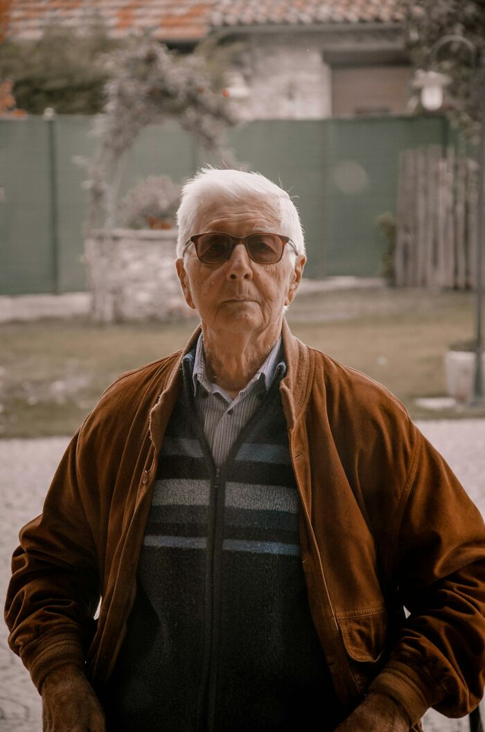 Elderly man wearing sunglasses and brown jacket standing outdoors, showcasing intelligence in a casual setting.