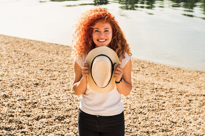 Woman with curly red hair holding a hat by the water, smiling happily in a moment people can't explain from psychic sessions