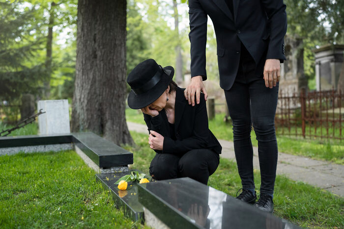 Woman in black outfit mourning at grave while another person comforts her during a psychic session outdoors