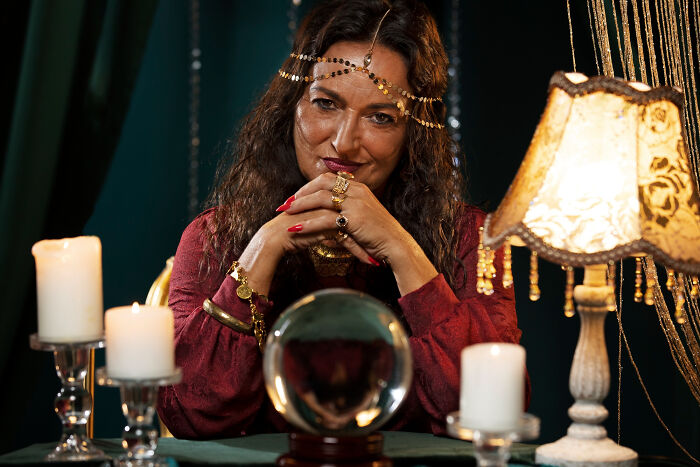 Woman psychic with crystal ball and candles wearing gold jewelry during a mysterious psychic session indoors