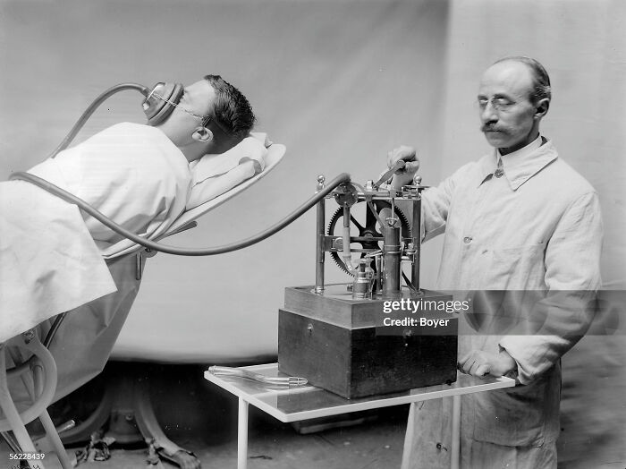 Vintage medical device with patient wearing a respirator mask connected to a mechanical apparatus operated by a healthcare worker.