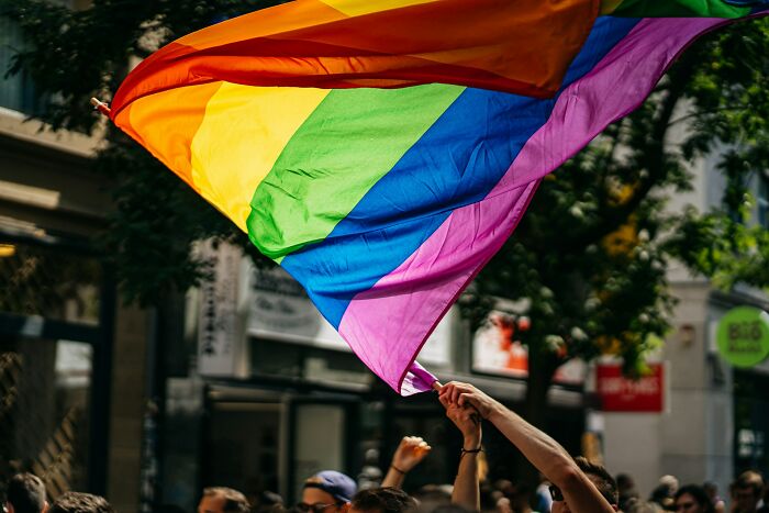 A person holding a large, colorful rainbow flag, with a blurred street and buildings in the background. Former priests and nuns experiences.