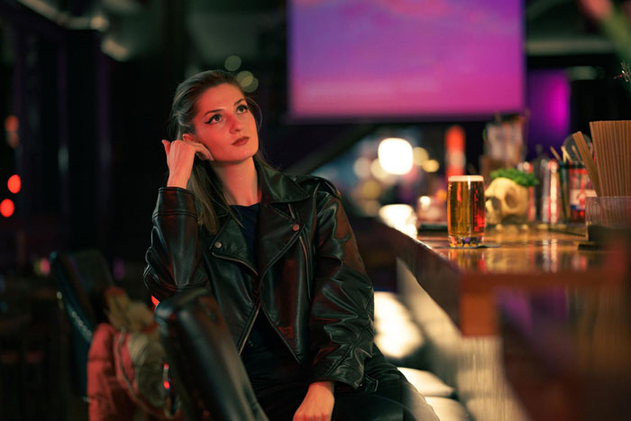 Young woman wearing a leather jacket sitting alone at a bar, reflecting on coping with ex-BF&rsquo;s trauma through comedy.