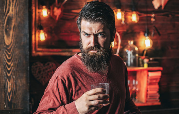 Bearded man in a dimly lit room holding a glass, symbolizing stalking after trauma and coping with comedy.