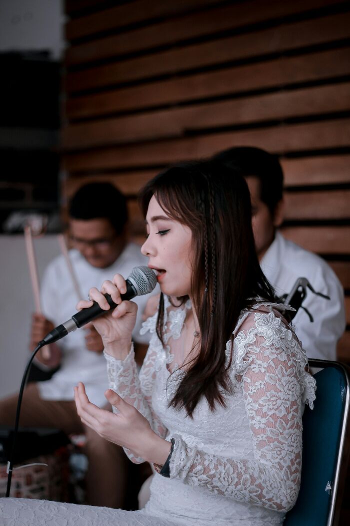 Bride in a lace dress singing into a microphone during a live performance at a wedding with a small band in the background.