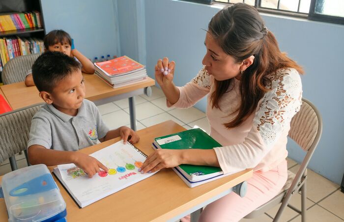 Teacher patiently helping young student with schoolwork in a classroom, illustrating kindness and good person qualities.