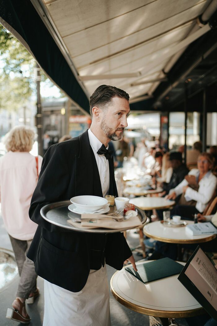 A waiter, in a bow tie and apron, carries a tray of dishes at an outdoor cafe. Some people rage quit their jobs here.
