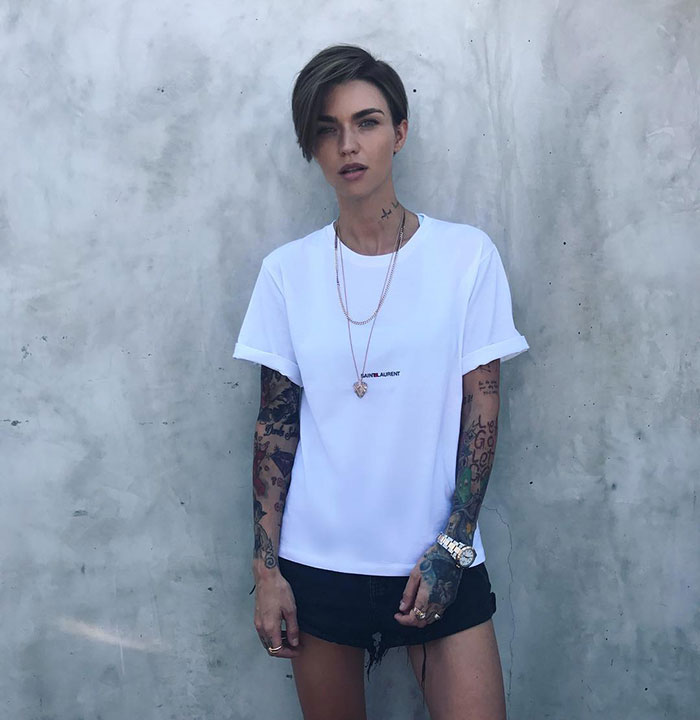 Person with tattoos wearing a white shirt and black shorts standing against a gray concrete wall after misconduct accusations. Person with tattoos wearing a white shirt and black shorts standing against a gray concrete wall after misconduct accusations.
