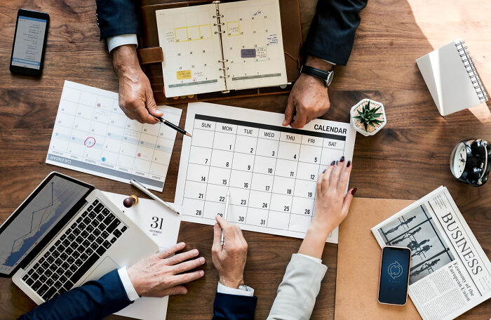 Three people, hands visible, plan on calendars at a wooden table, amidst work tools. Absurd rules that tanked miserably.