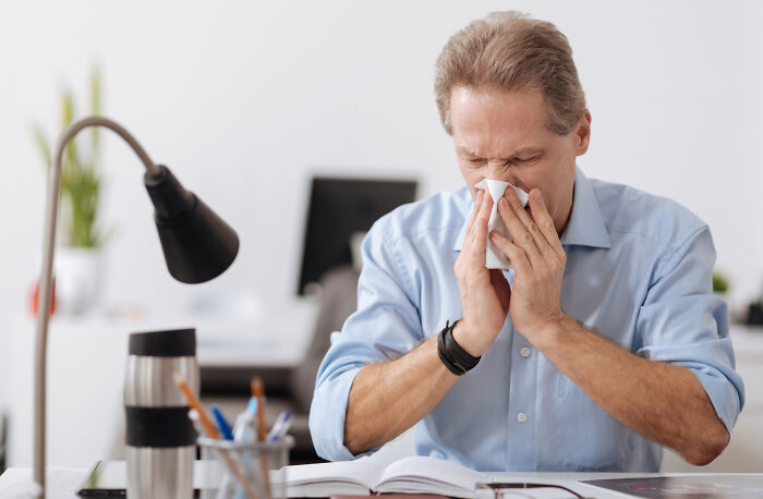 A man in a blue shirt is sneezing into a tissue at his desk, highlighting absurd rules in schools and workplaces.