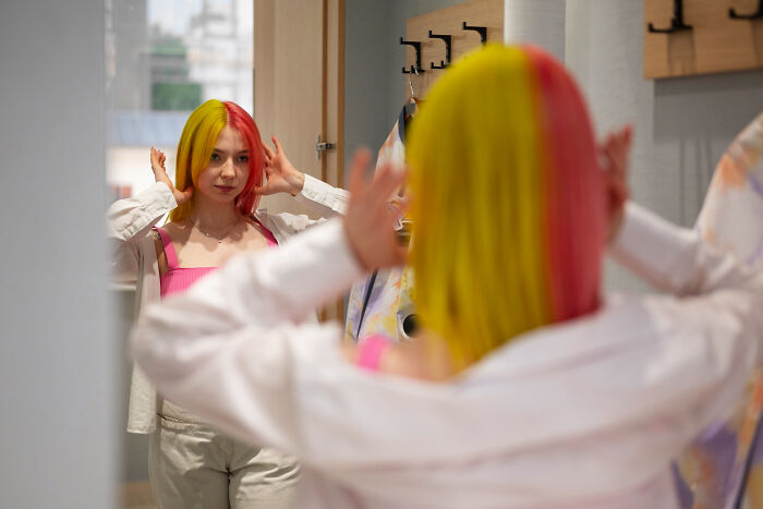 A young woman with half pink, half yellow hair looks in a mirror. Her reflection shows her considering absurd rules.