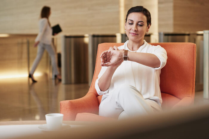 A woman in white checks her watch while seated in an orange chair, reflecting on absurd rules in schools and workplaces.