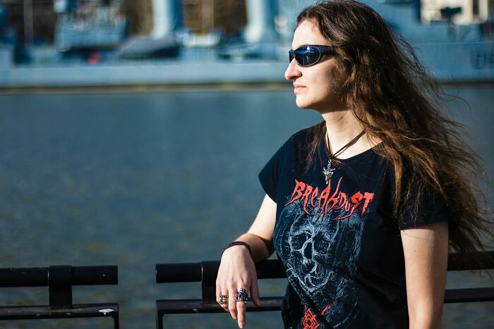 Woman in sunglasses and a black band t-shirt, long hair blowing, stands by water. Absurd rules fail.