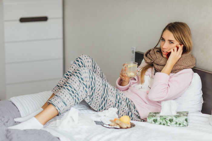 A sick woman in bed, wrapped in a scarf, drinking tea and on the phone, reflecting absurd rules and their failure.