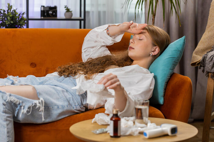 A sick young woman with long curly hair rests on an orange couch, hand to her forehead, surrounded by tissues and medicine, highlighting absurd rules.