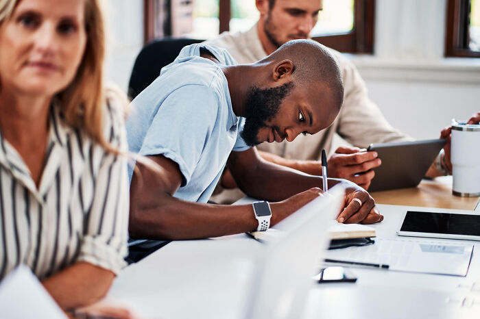 Diverse colleagues in a meeting, with one man taking notes. This scene highlights the absurd rules schools and workplaces implement.