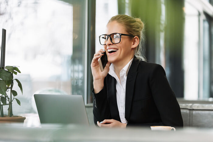 Smiling woman in glasses talking on phone, next to laptop and coffee. Discussing absurd rules that schools and workplaces tried to implement.