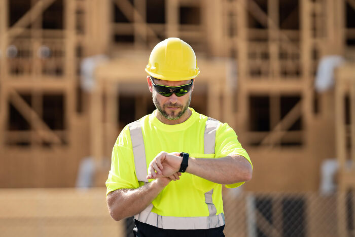 A construction worker in a yellow hard hat and safety vest checks his smartwatch, frustrated with absurd rules.