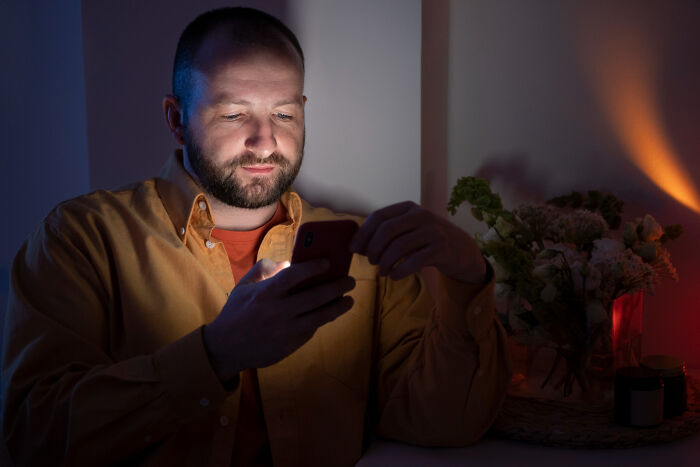 A man in a yellow shirt uses his glowing phone in a dim room. He's smiling, reflecting on absurd rules.