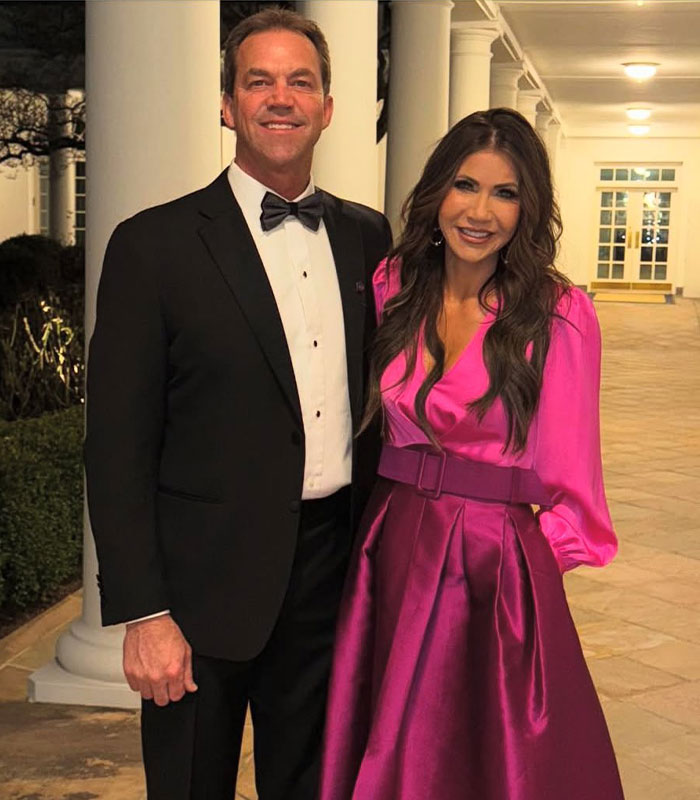 Bryan Noem in a black tuxedo and a woman in a bright pink dress posing together indoors on a tiled floor.