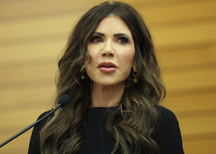 Kristi Noem speaking at a podium with microphone, as internet slams SNL for mocking her husband Bryan over scandal.