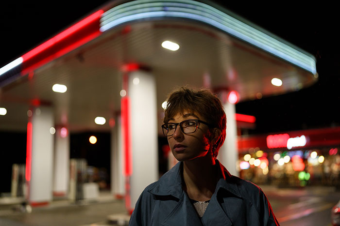 A woman with short hair and glasses at a gas station at night, illuminated by red and white lights, creating a scary atmosphere.