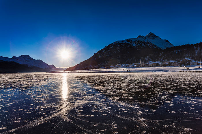 Sunlight glares across a frozen lake with ice skaters, snow-covered mountains, and a village. Scary Experiences People Wish To Forget.