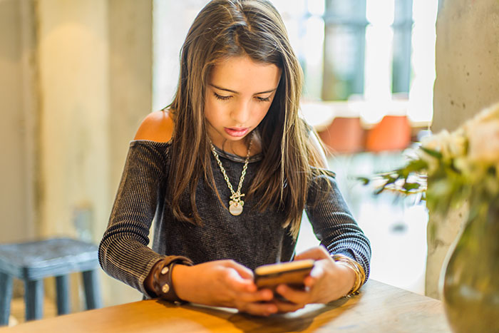 A young girl with long brown hair, wearing a gray shirt and necklace, looking intently at her phone. She is having a scary experience.