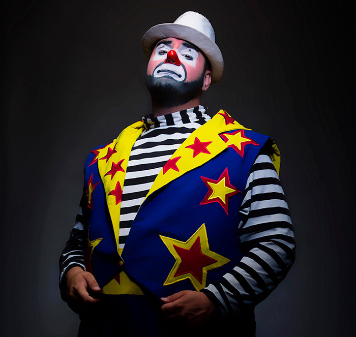 A sad clown in a white hat, striped shirt, and blue vest with stars, looking up, evoking scary experiences.