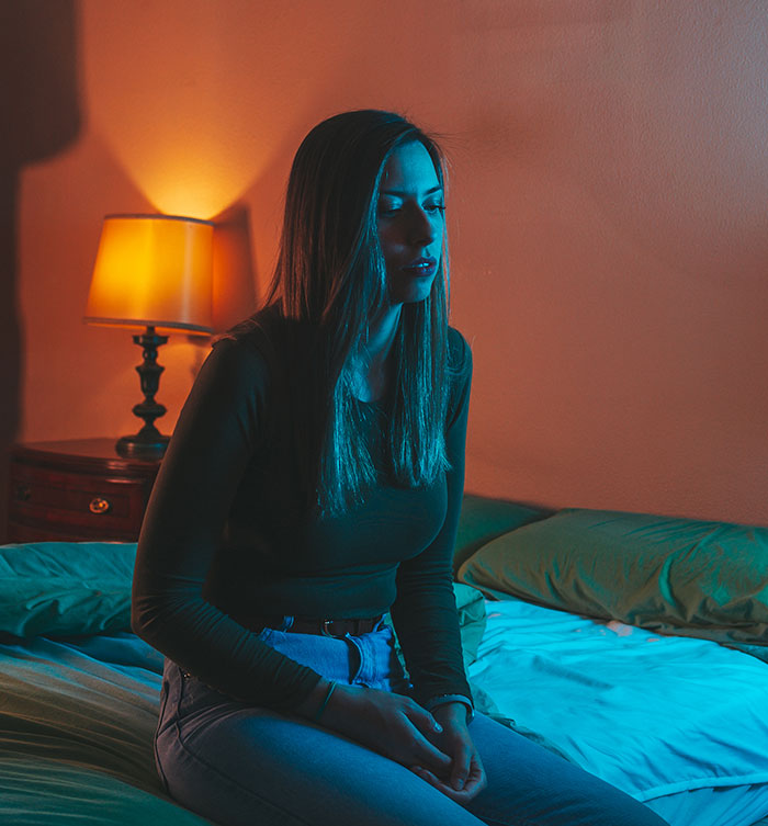 A young woman sits on a bed, illuminated by blue light with an orange lamp in the background, depicting scary experiences.