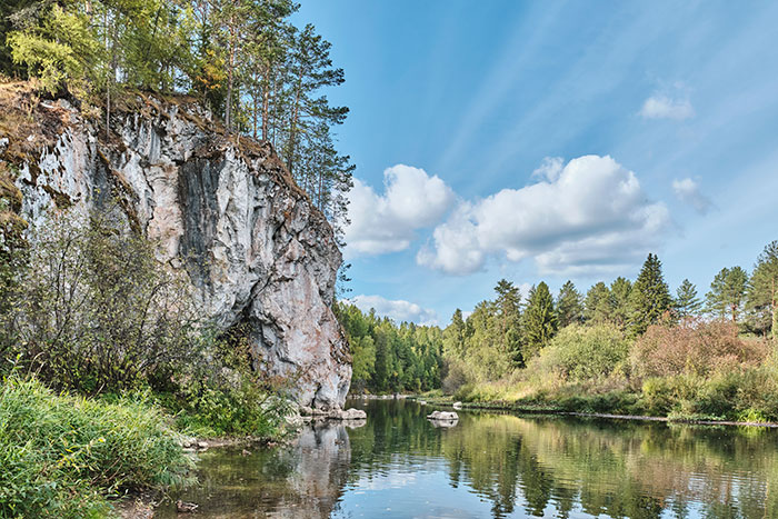 A serene river flows past a towering cliff covered in trees under a partly cloudy sky, a view that makes you wish to forget your scary experiences.