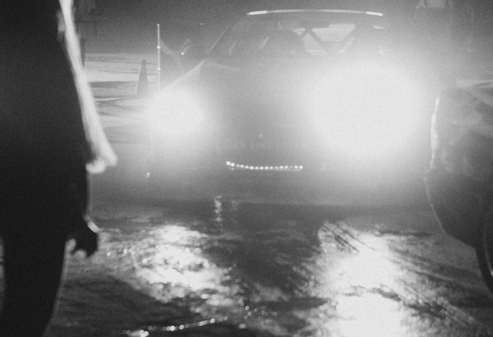 Black and white photo of a car with bright headlights on a wet road at night, creating a scary experience.