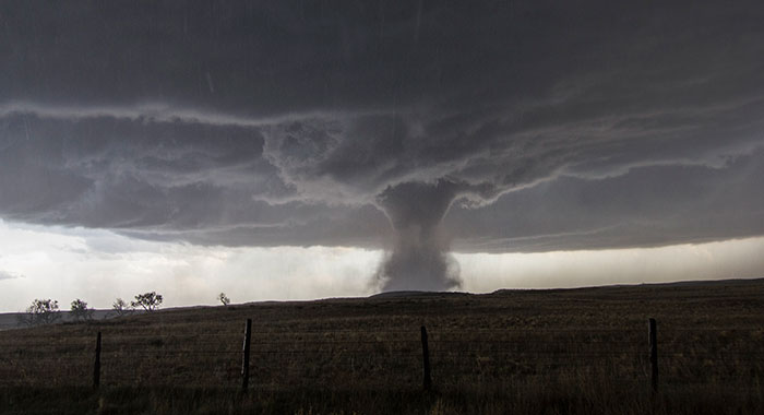A scary experience: a powerful tornado touching down in a rural field under a dark, stormy sky, seen through a fence.